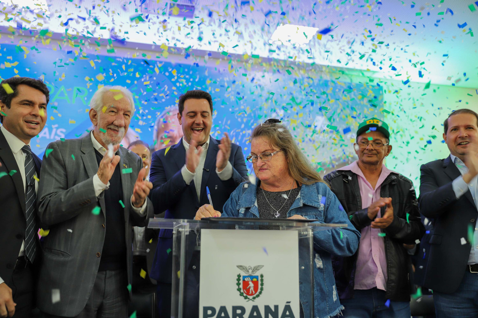 Seis pessoas em um palco comemorando, com uma pessoa idosa assinando em um púlpito.