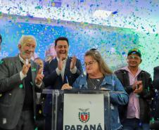 Seis pessoas em um palco comemorando, com uma pessoa idosa assinando em um púlpito.