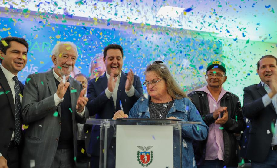 Seis pessoas em um palco comemorando, com uma pessoa idosa assinando em um púlpito.