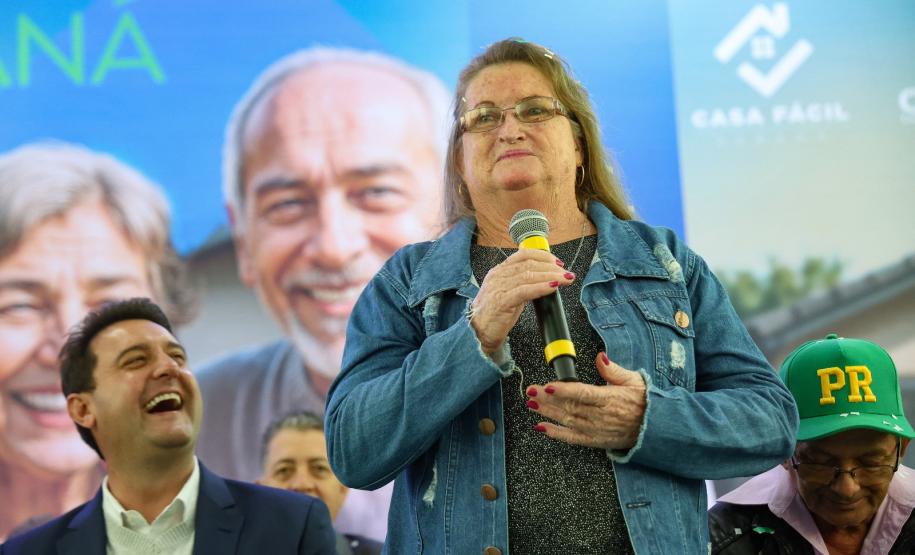 Três pessoas em um palco, com uma pessoa idosa falando ao microfone.