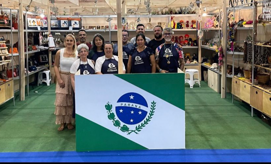 A foto mostra um grupo de expositores em um estande da Fenearte 2025, representando o estado do Paraná. Eles estão reunidos atrás de um painel com a bandeira do estado. Ao fundo, é possível ver prateleiras com peças de artesanato como bolsas, bonecas, esculturas e bijuterias, organizadas de forma colorida e atraente. O estande tem estrutura de madeira clara, piso verde e ambiente iluminado.